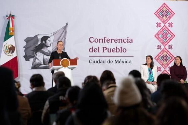 Presidenta Claudia Sheinbaum habla frente a un atril con el escudo nacional, mientras se proyectan gráficas de crecimiento turístico en una pantalla.