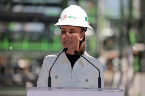 Presidenta Sheinbaum con casco blanco habla frente a torres de destilación de la Refinería Tula, trabajadores escuchan.