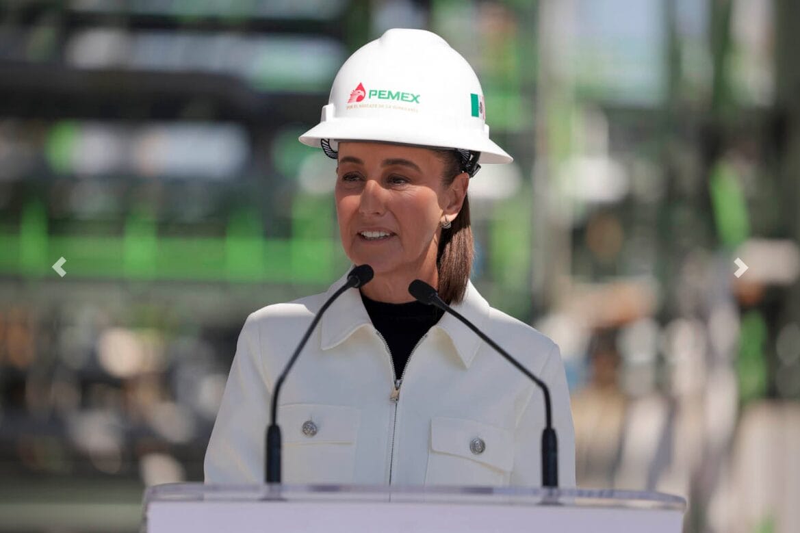 Presidenta Sheinbaum con casco blanco habla frente a torres de destilación de la Refinería Tula, trabajadores escuchan.